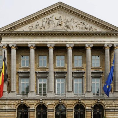 Facade of a historic building featuring tall columns, Belgium and European flags, and intricate sculptures atop the pediment.