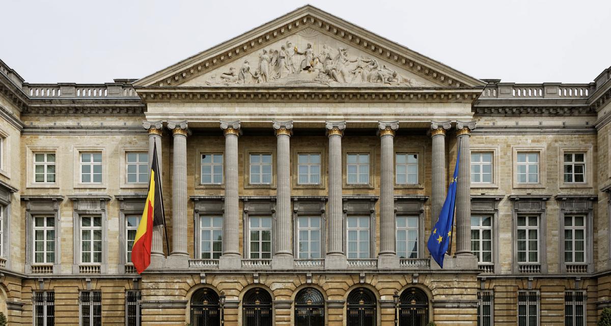 Facade of a historic building featuring tall columns, Belgium and European flags, and intricate sculptures atop the pediment.