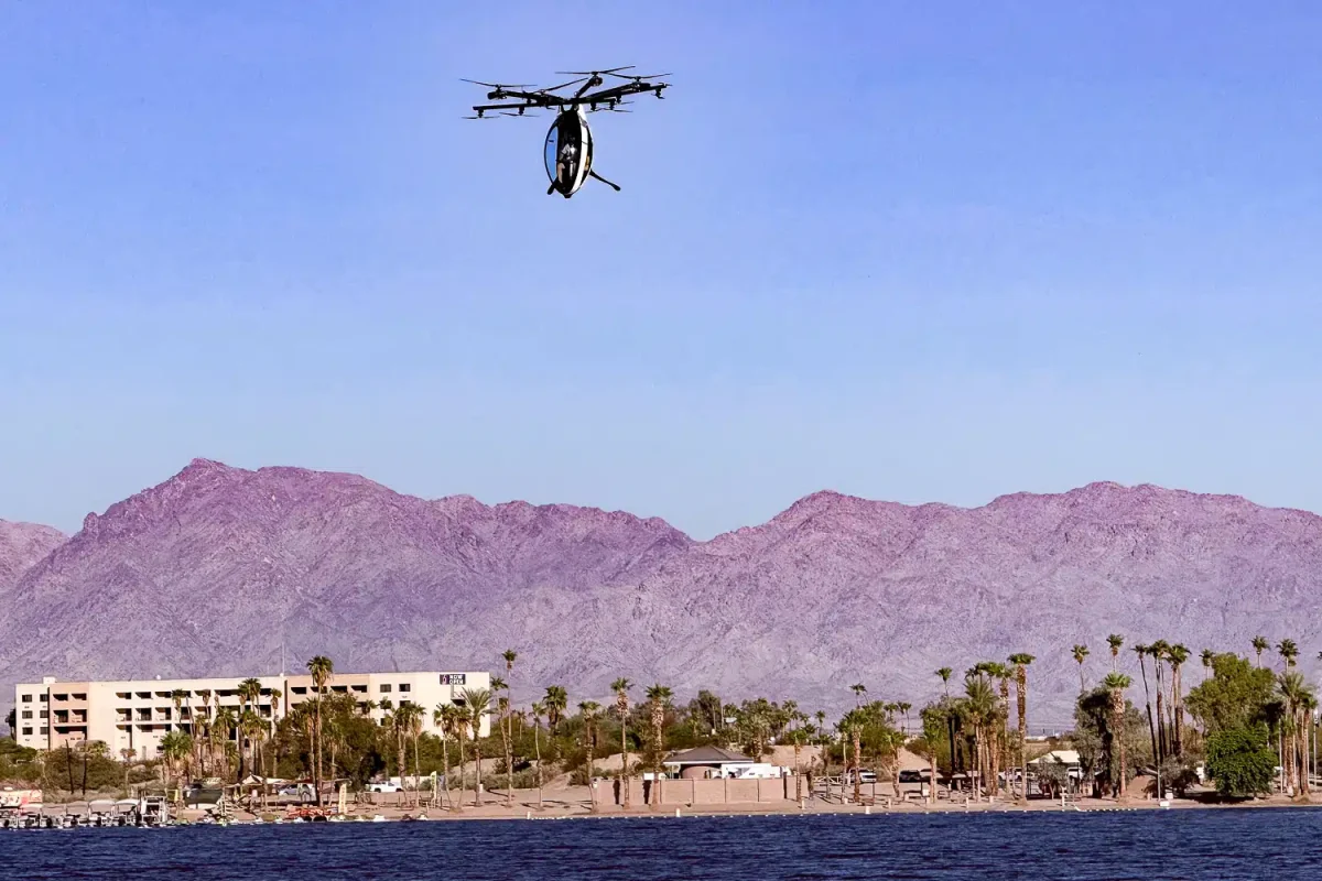 A helicopter hovers above a lakeside resort with palm trees and sandy beaches, framed by purple mountains under a clear blue sky.