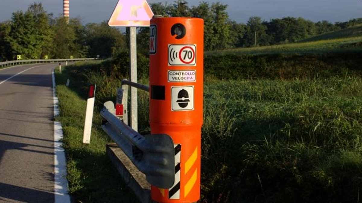 A roadside speed camera in an orange housing displays a 70 km/h speed limit sign, surrounded by greenery and safety barriers.