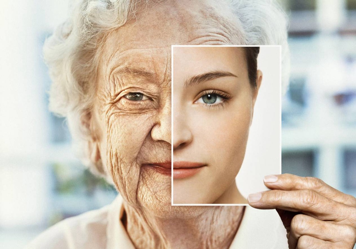 An elderly woman holds a photo of a young woman's face over half of her own, contrasting aged skin with youthful features.