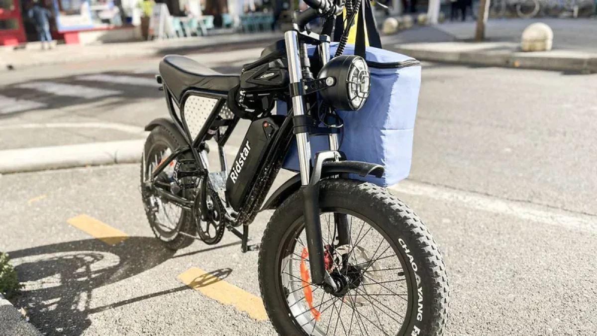 A sleek electric bike with a black frame and orange accents parked on a city street, featuring a blue bag attached to the handlebars.
